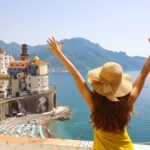 Summer holiday in Italy. Back view of young woman with straw hat and yellow dress with raised arms looking at Atrani village, Amalfi Coast, Italy